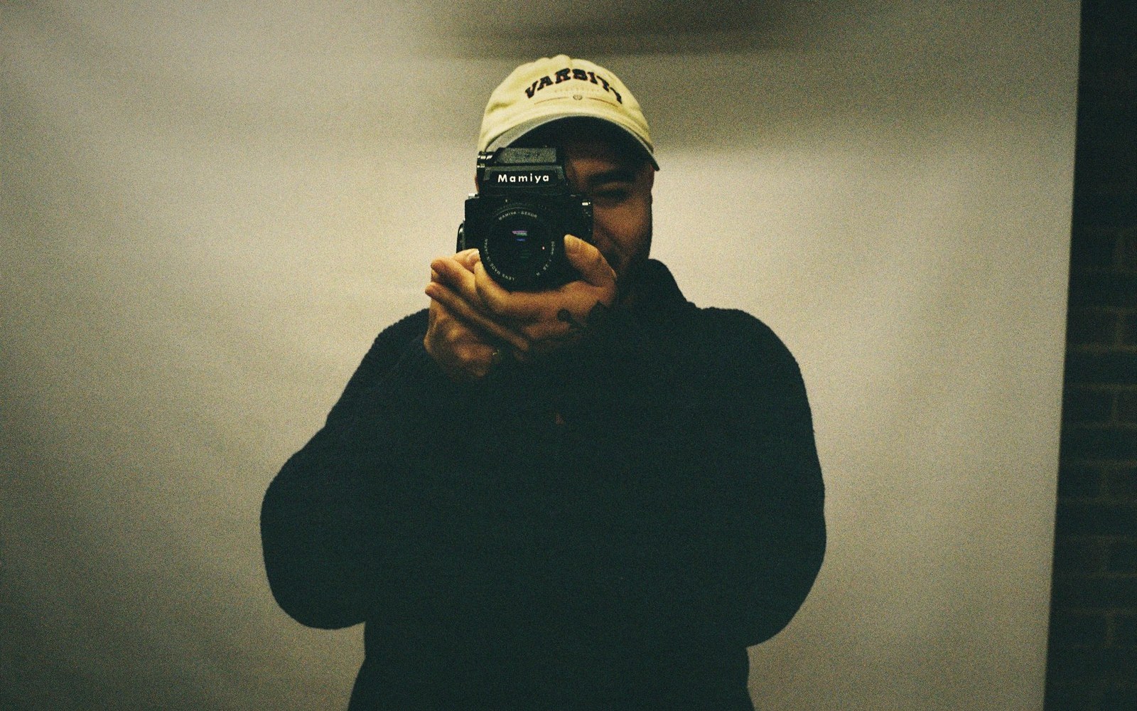Man in a varsity cap holds a Mamiya medium-format camera up to his face
