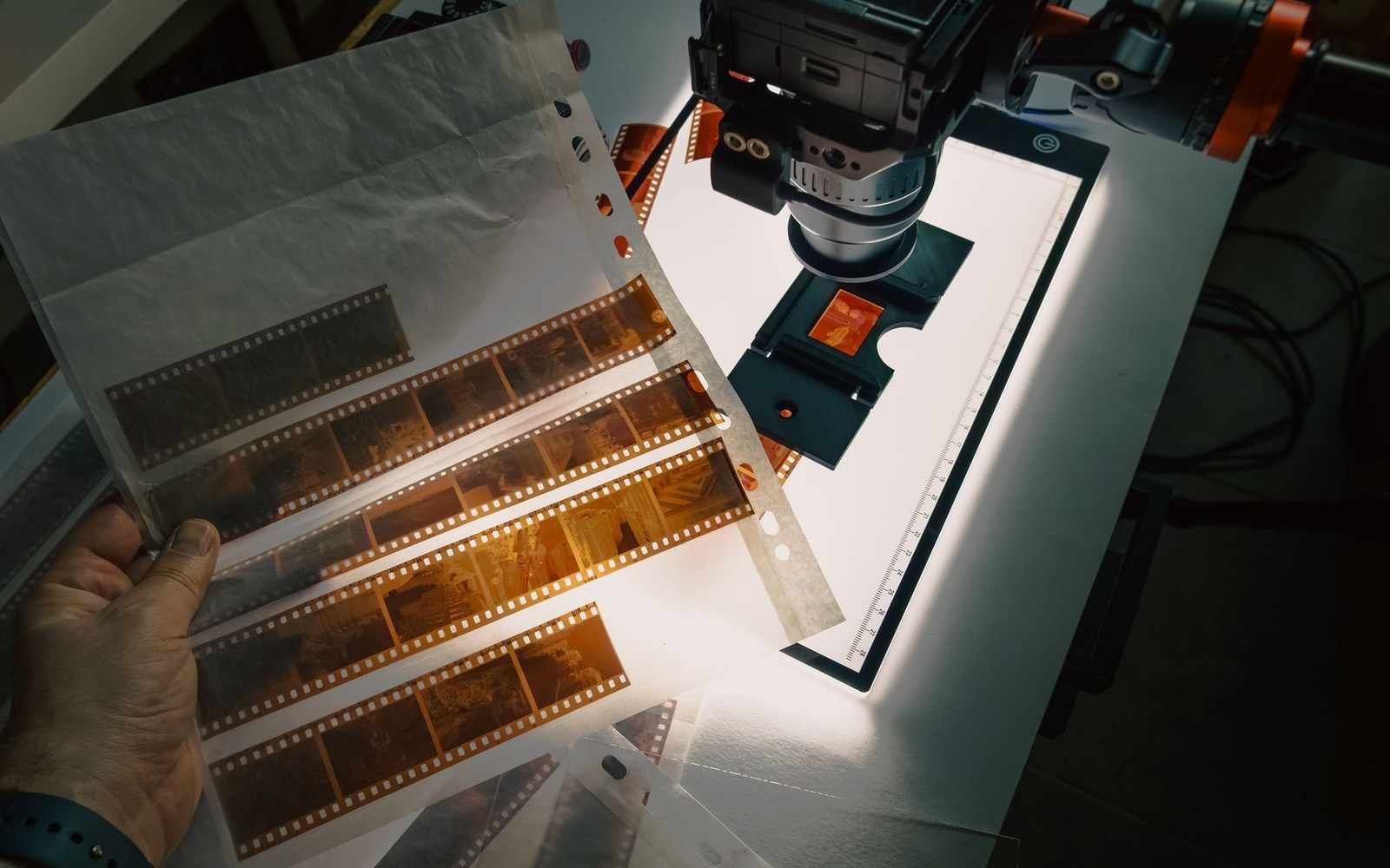 A hand holds negative film strips beside a camera setup on a lightbox for digitizing